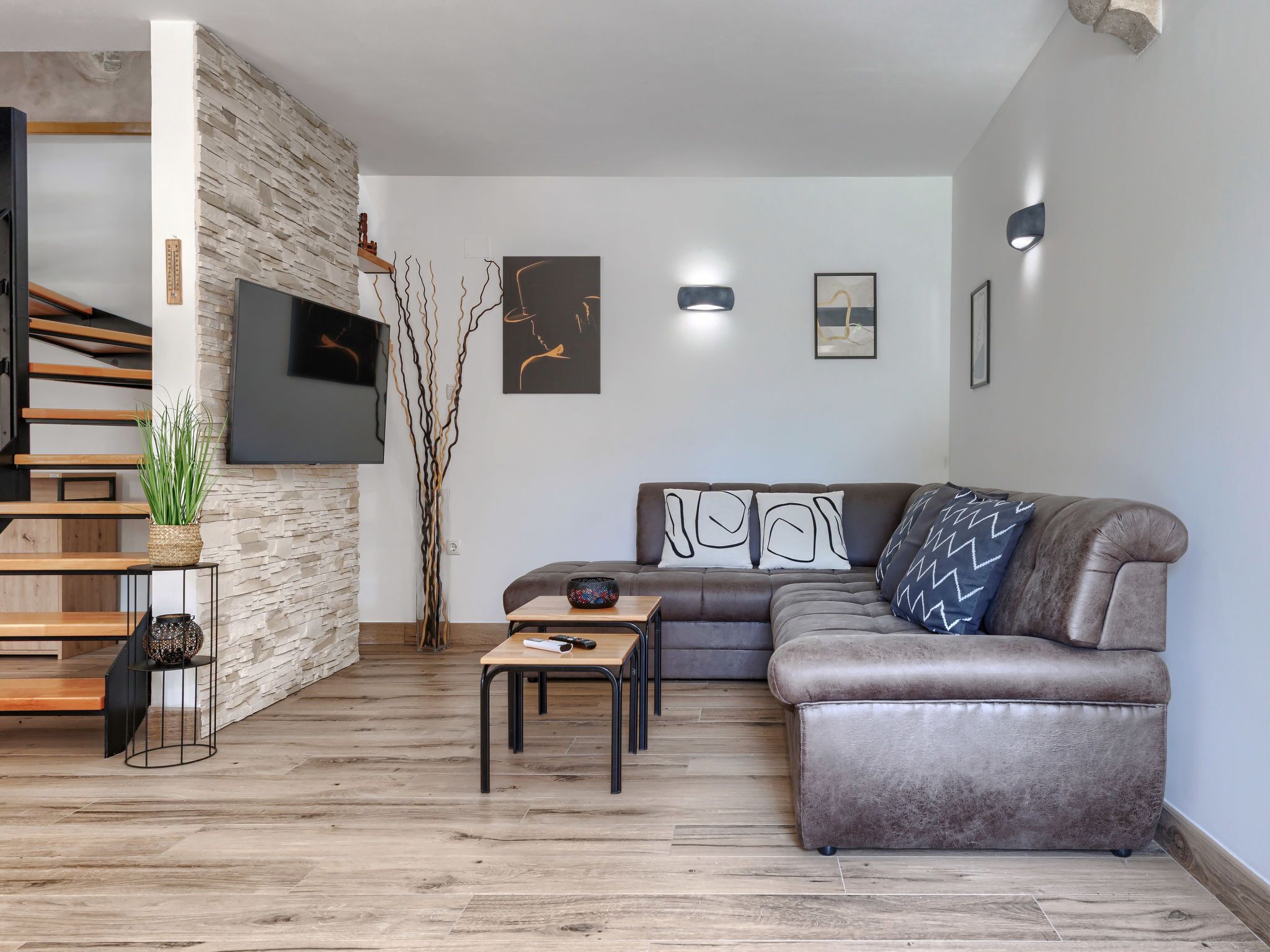 A modern living room with a stone accent wall, a grey leather sofa, a wooden coffee table, and a staircase with wooden steps.