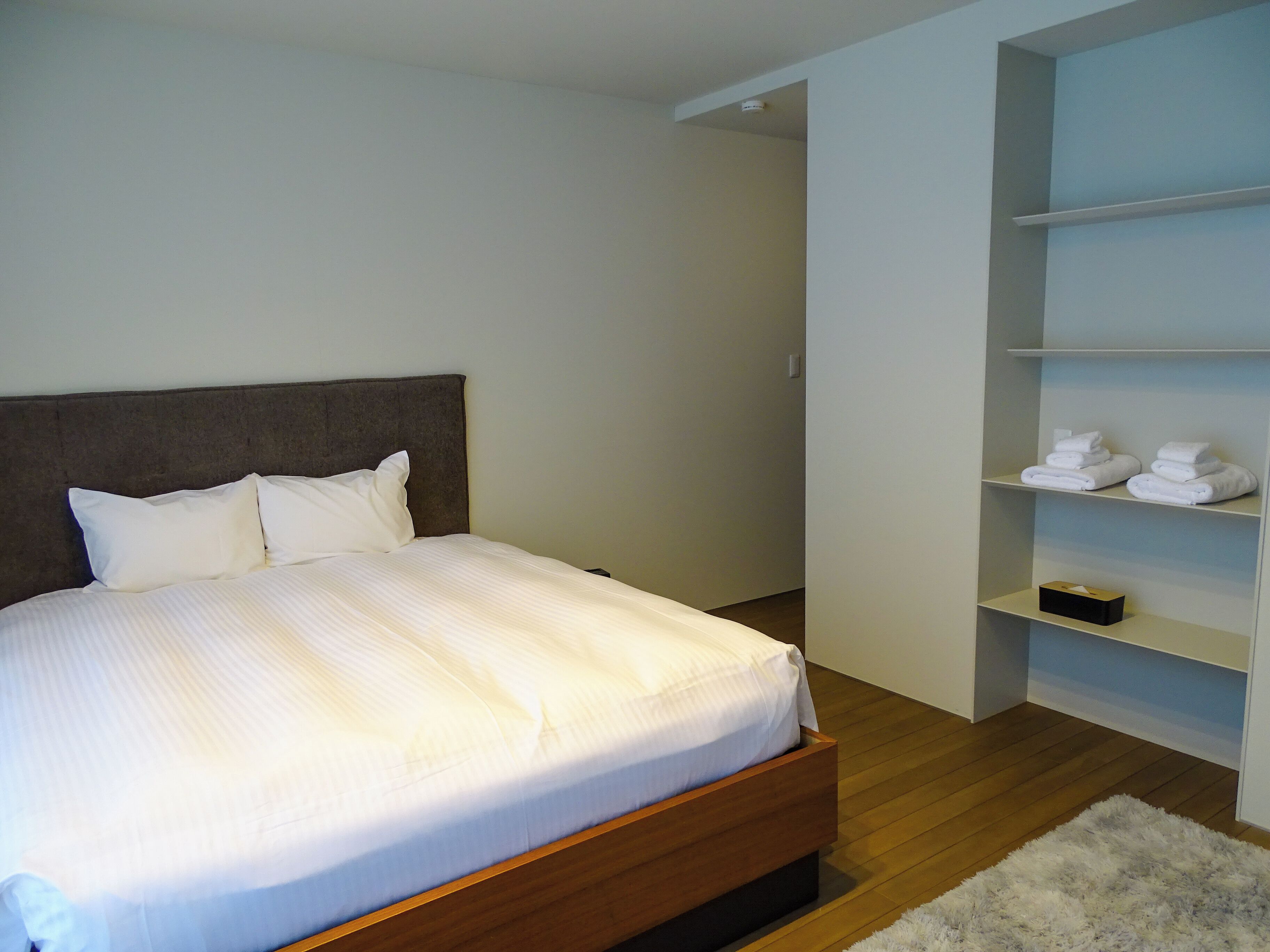 A neatly made bed with a wooden headboard, a white wall, and a built-in shelf with folded towels.