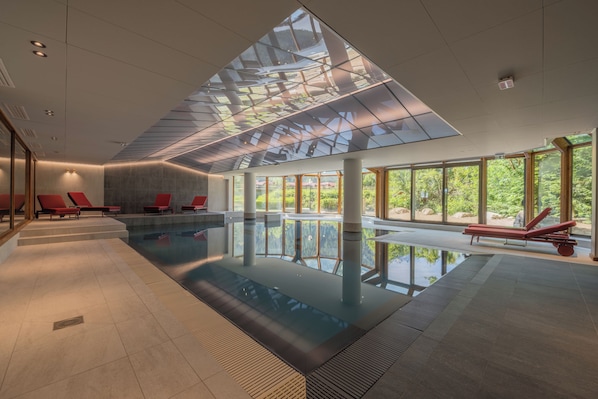 A modern indoor swimming pool with clear glass ceiling, red lounge chairs, and large windows.