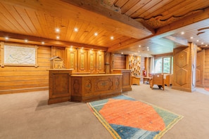A wooden interior with a central wooden counter, bookshelves, and a large rug.