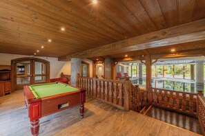 A wooden pool table in a room with a wooden ceiling and a balcony with a railing.