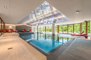 A modern indoor swimming pool with clear glass ceiling, red lounge chairs, and large windows offering a view of greenery.