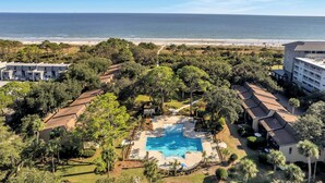 Vista aérea de un complejo turístico con piscina rodeada de exuberante vegetación, una playa al fondo y varios edificios.