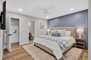 Un dormitorio con una cama, dos mesitas de noche, un ventilador de techo y una puerta que da al baño.