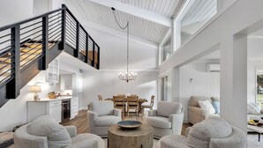 A spacious living area with a high ceiling, a staircase with black railings, and a chandelier. There are several grey armchairs and a round coffee table with a bowl on it.