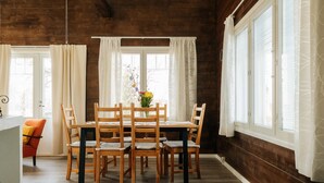 A dining area with a wooden table and chairs, a vase with flowers, and large windows with curtains.