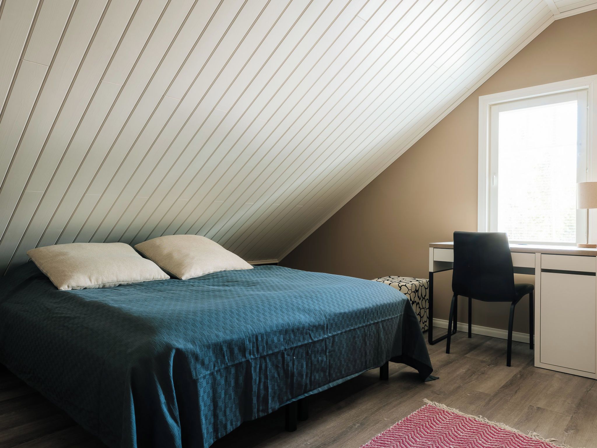 Un dormitorio con techo inclinado, una cama con colcha azul, dos almohadas, un escritorio con silla y una ventana.