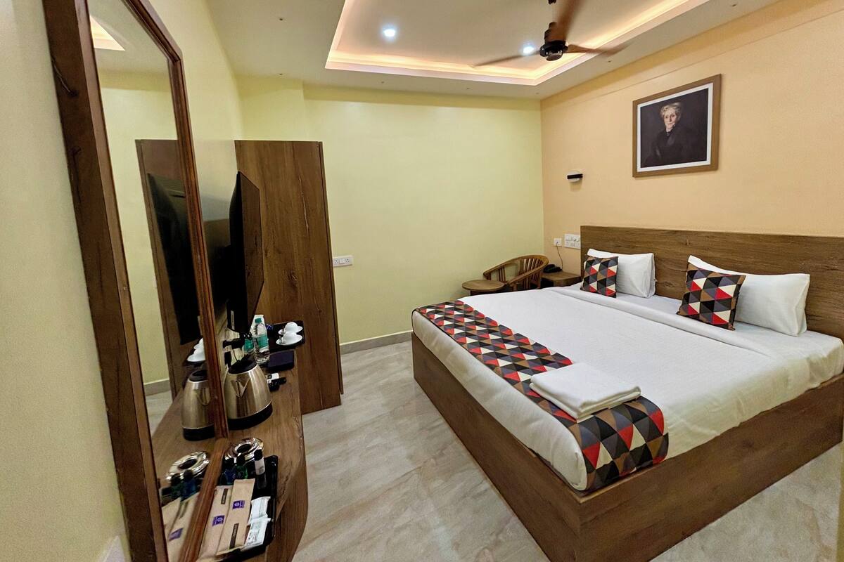A hotel room with a large bed, a wooden wardrobe, a desk with a kettle, and a ceiling fan.
