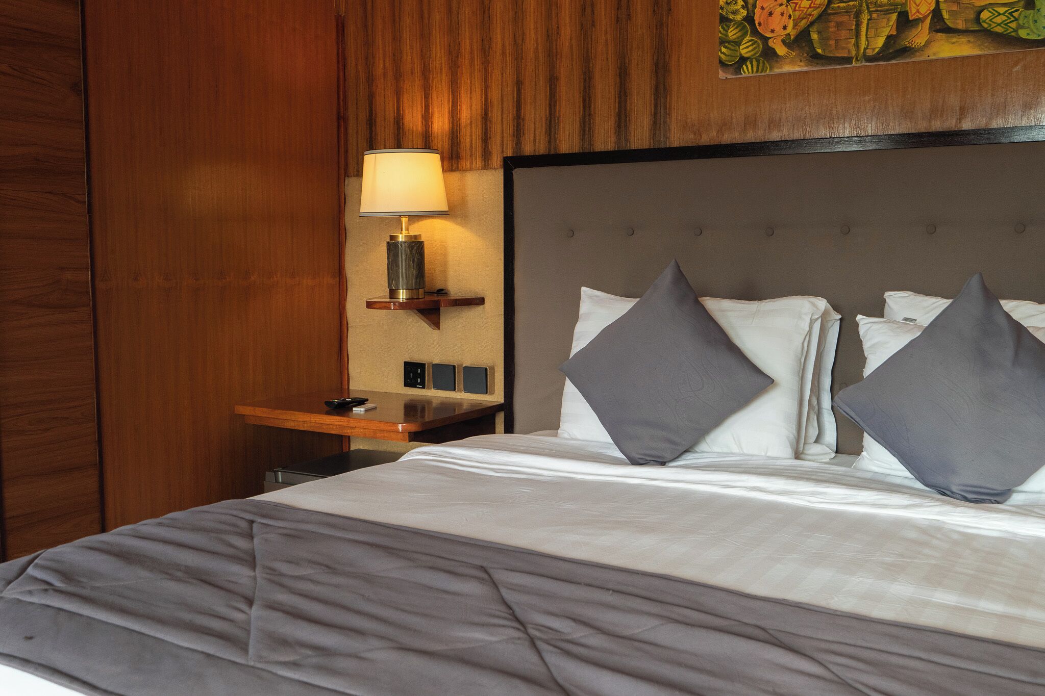 Una cama bien hecha con ropa de cama gris y dos almohadas grises, una mesita de noche con una lámpara y un panel de pared de madera.