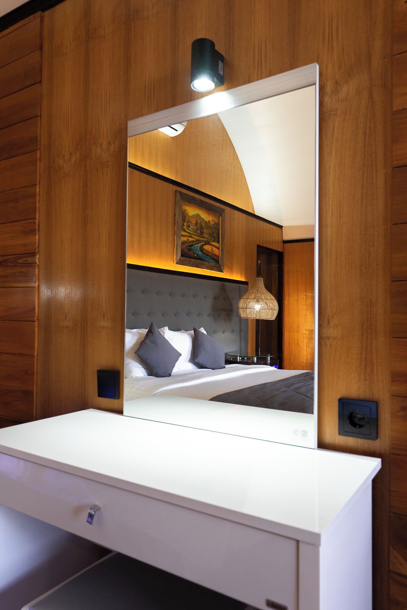 A modern hotel room with a large mirror, wooden paneling, a tufted headboard, a bedside table, and a hanging light fixture.