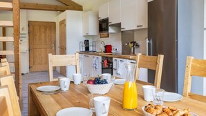 Une cuisine moderne avec des poutres en bois, des meubles blancs, un réfrigérateur en acier inoxydable et un coin repas avec une table en bois mise pour le petit-déjeuner.