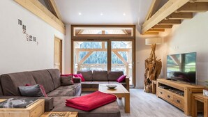 Un salon moderne avec un canapé d’angle, une table basse en bois et un téléviseur posé sur un meuble en bois. De grandes fenêtres offrent une vue sur l’extérieur.