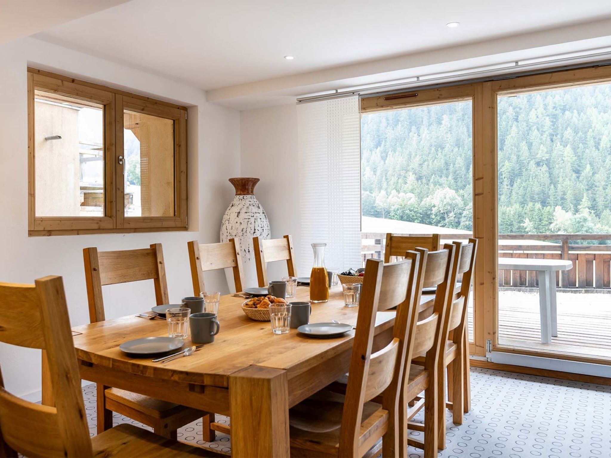 Un comedor con una mesa y sillas de madera, un jarrón y vistas a un bosque a través de amplios ventanales.