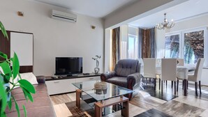 Een moderne woonkamer met een flatscreen televisie, comfortabele zithoek, een glazen salontafel en een eetgedeelte met witte stoelen.