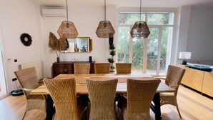 A dining area with a wooden table and wicker chairs, a mirror, and a view of greenery through large windows. - Oasis in Roche. Design villa (Roche)