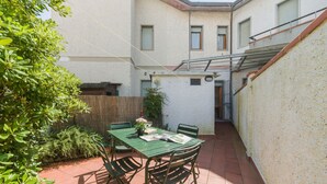Eine kleine Terrasse mit Tisch und Stühlen, umgeben von einem weißen Gebäude mit Holzzaun und Grünpflanzen.