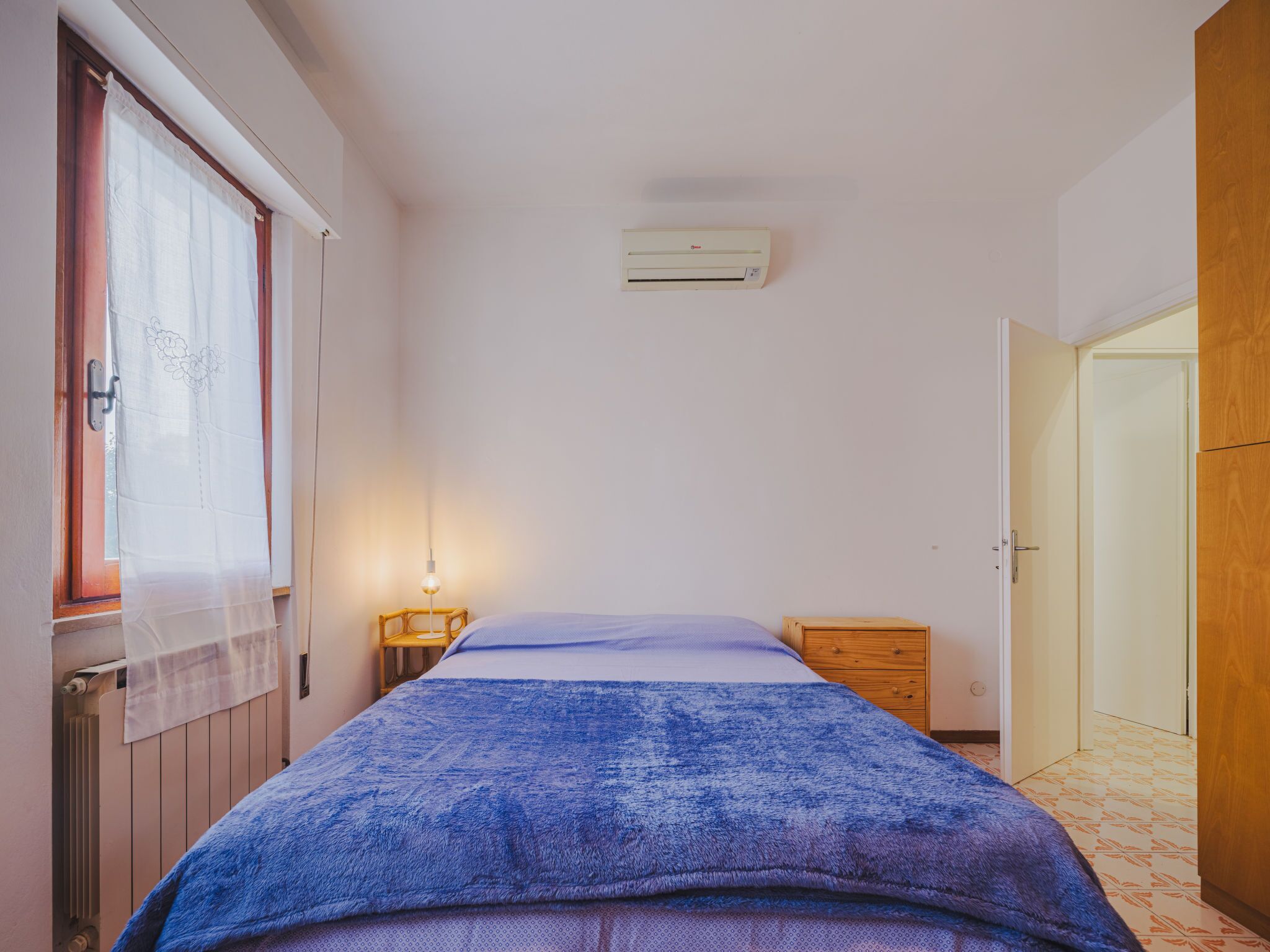 A bedroom with a bed, a bedside table, a nightstand, a window with curtains, an air conditioning unit, and a door.