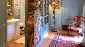 A room with a wooden floor, a patterned rug, a chair, a lamp, and a door with a mirror. - Le Jardin Perché: Suite for Two (MOULEYDIER)