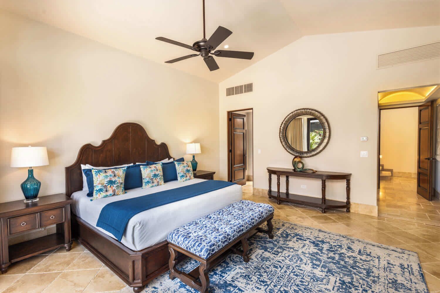 Une chambre à coucher avec un lit en bois, deux tables de chevet, un ventilateur de plafond, un grand miroir et un tapis à motifs.
