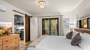 A bedroom with a large bed, a wooden dresser, a TV, a kitchen area, and a sliding glass door leading to a balcony with a view of trees. - Stylish Oak Creek Home w/ 2 Suites, Spa & Firepit (Sedona)