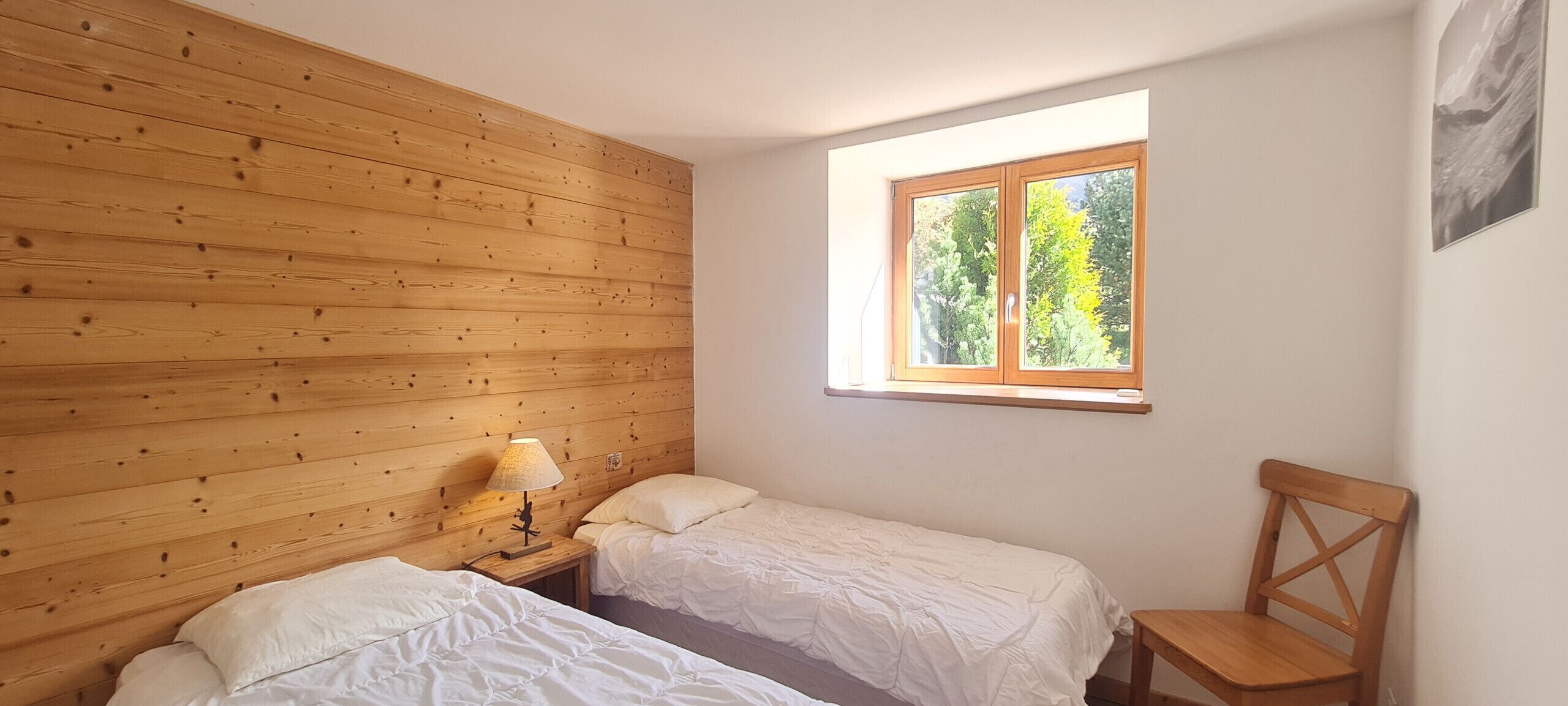 A room with two single beds, a wooden headboard, a small table with a lamp, and a window with a view of trees.
