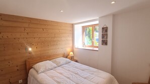 A bedroom with a wooden headboard, a bed with white bedding, a nightstand with a lamp, and a window with a view of greenery.