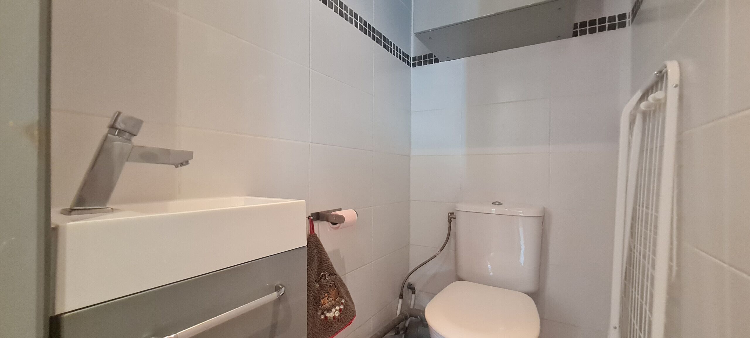 A small, clean bathroom with white tiles, a toilet, a sink with a faucet, and a towel hanging on the wall.