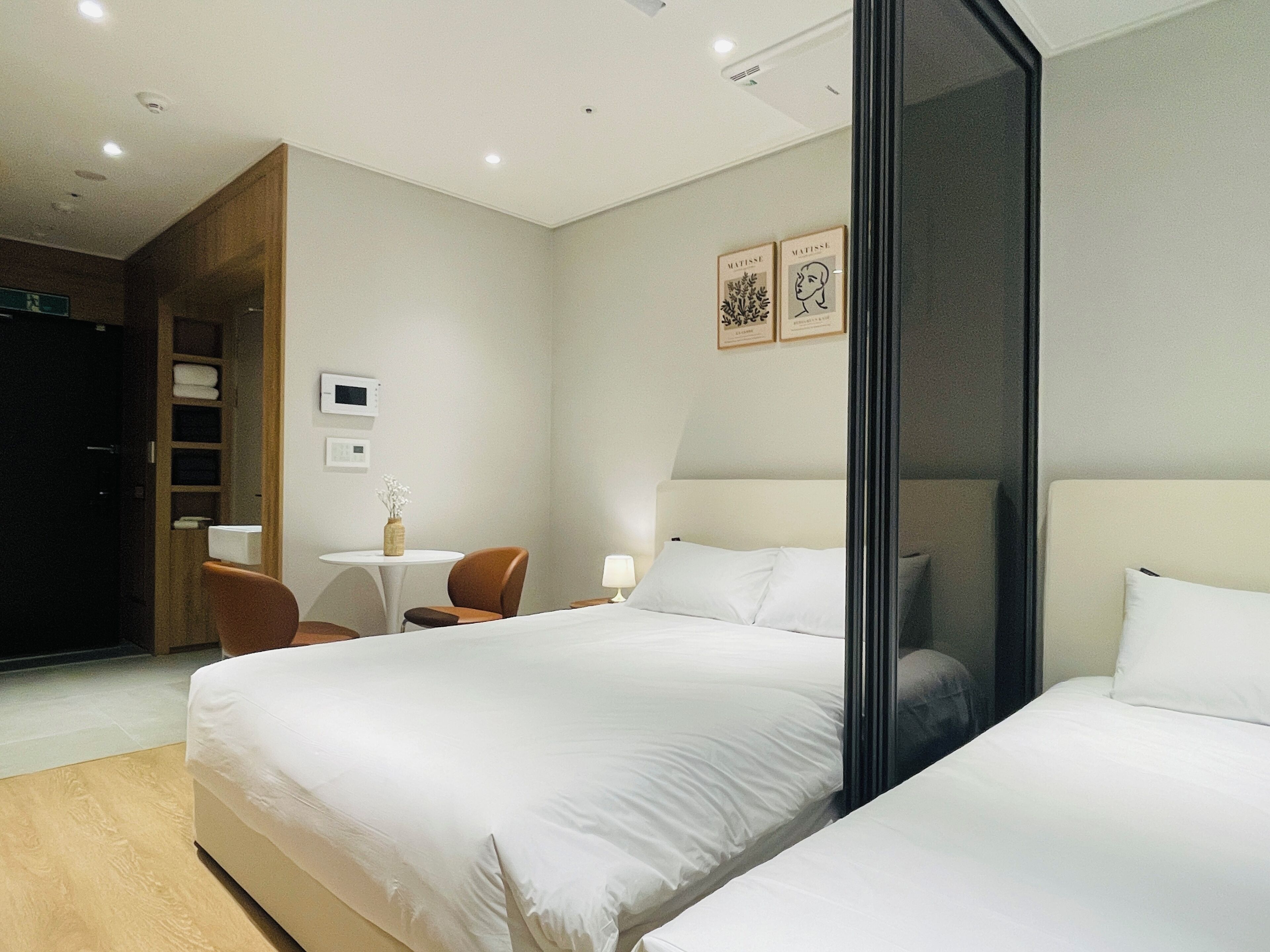 A hotel room with a large bed, a smaller bed, a desk with a chair, a small table with a vase, and two framed pictures on the wall.