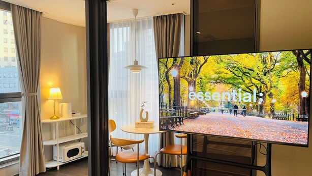 A modern hotel room with a large TV displaying a park scene, a white desk with a lamp, a chair, and a small shelf with books. - villiv Inn The City Namsan (Seoul)