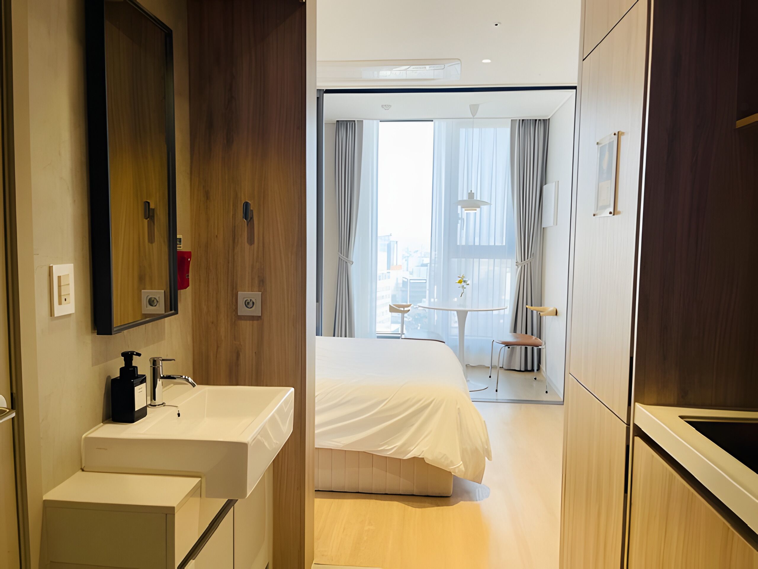 Une chambre d’hôtel avec une armoire en bois, un lavabo blanc, un lit recouvert d’une couverture blanche, une petite table avec une chaise et une grande fenêtre avec des rideaux.