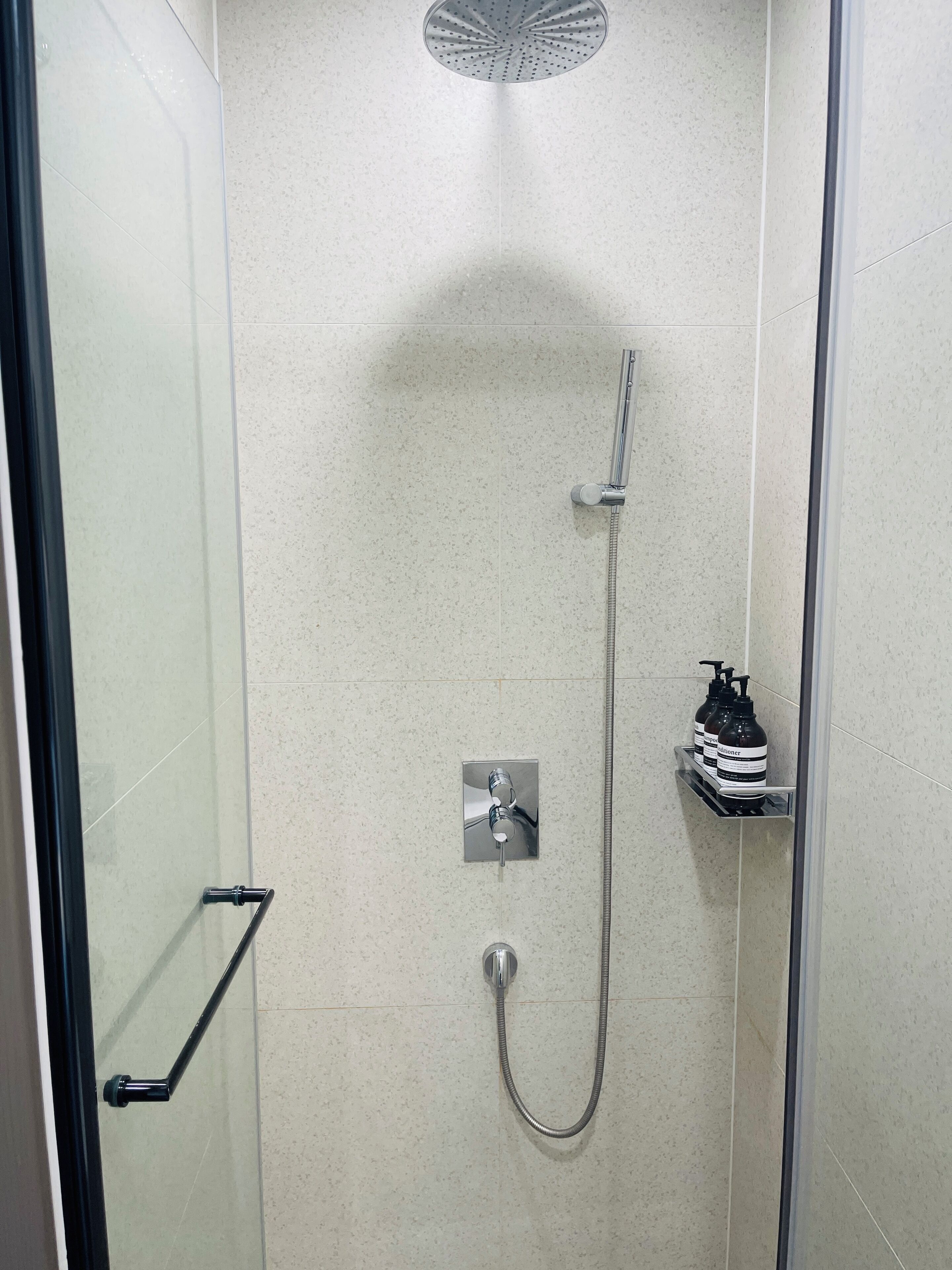 A modern shower with a handheld showerhead and a fixed showerhead, a glass door, and a small shelf with toiletries.