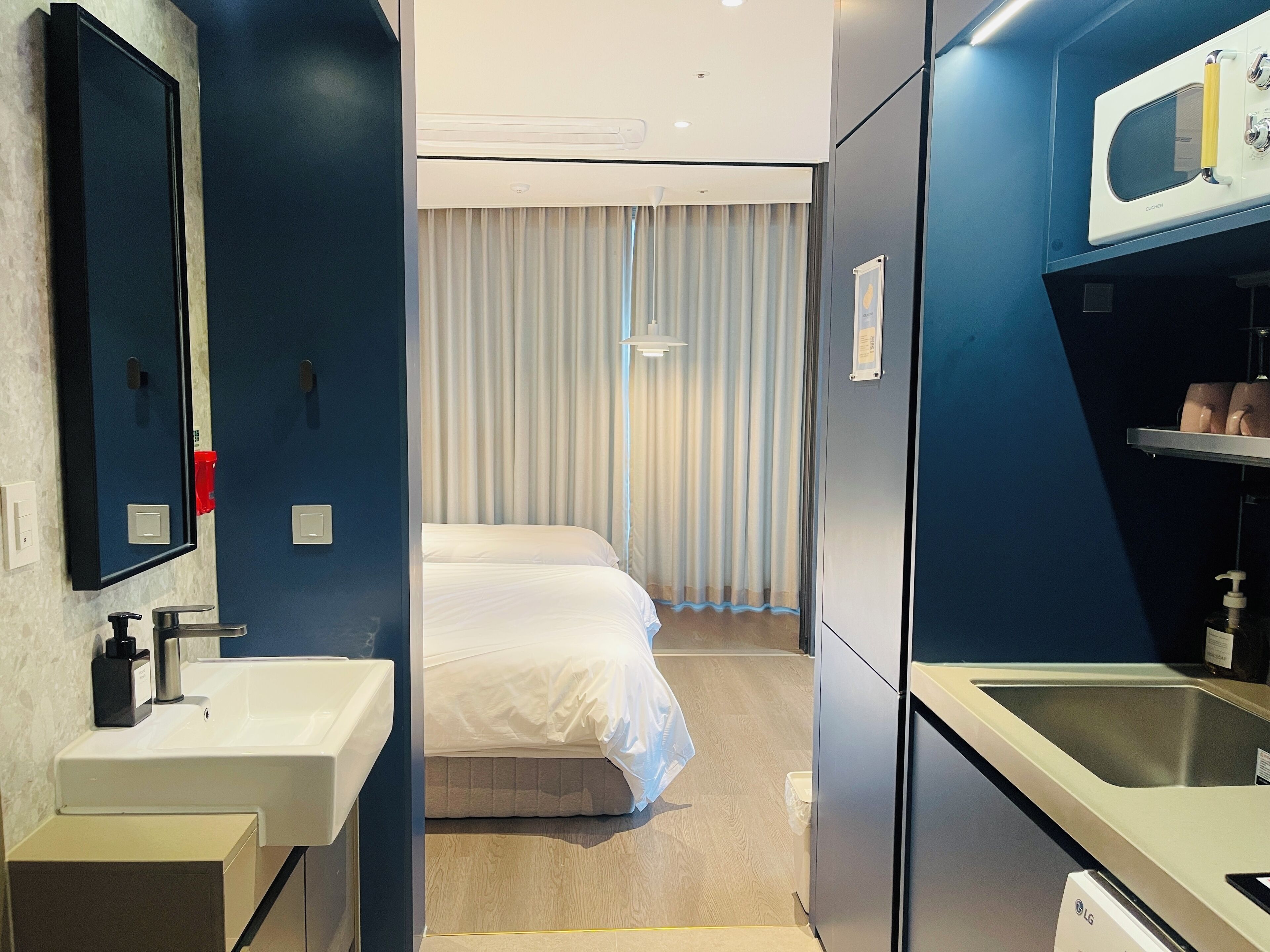 Quarto de hotel com cama, banheiro com pia e espelho, e uma pequena cozinha.