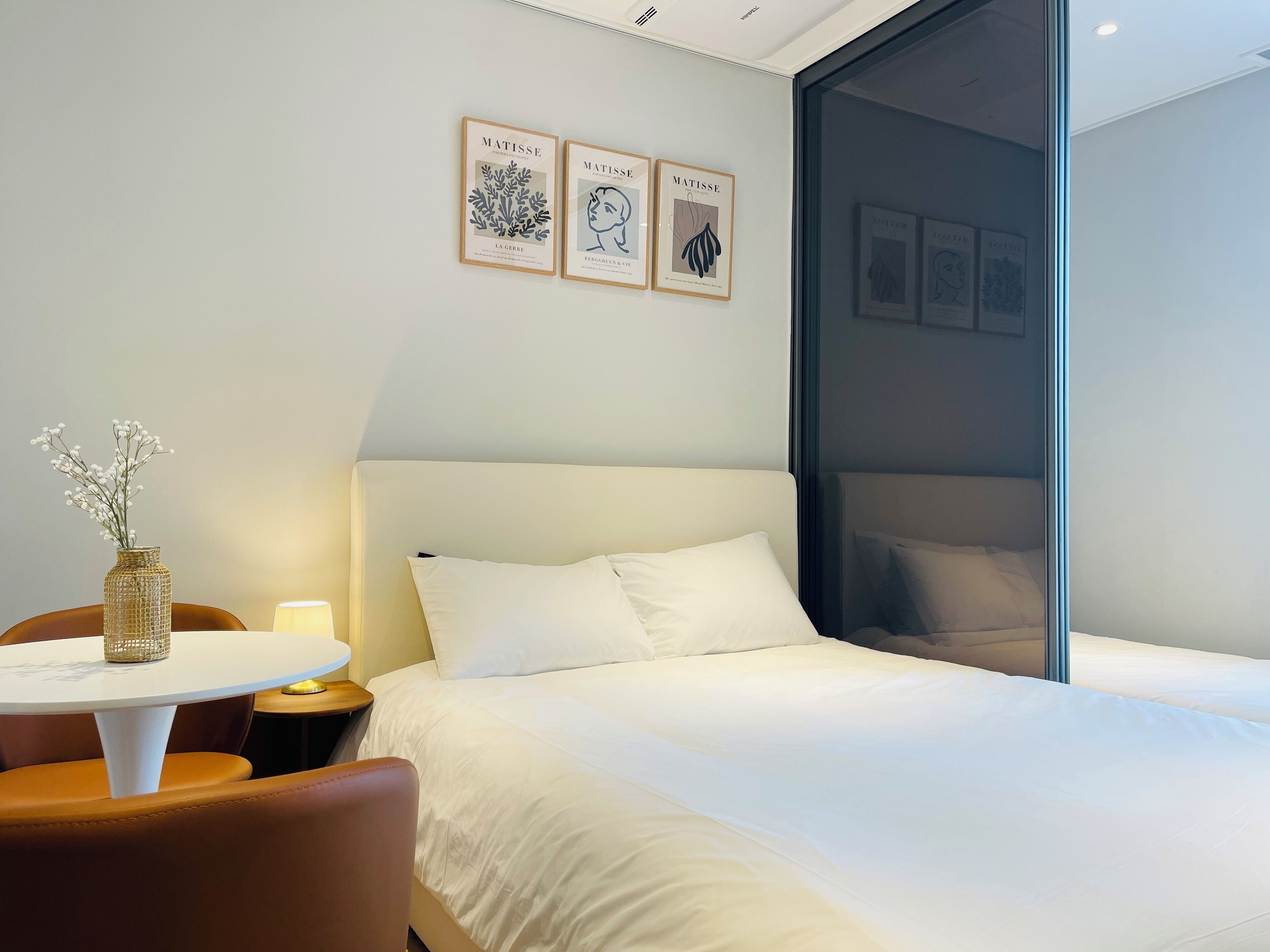 A hotel room with a bed, a round table with a vase, and three framed artworks on the wall.