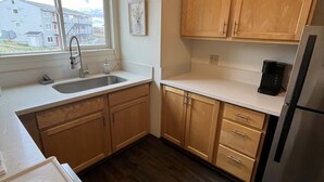A compact kitchen with wooden cabinets, a stainless steel refrigerator, and a window with a view of buildings.