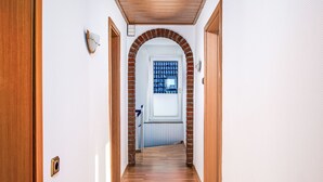 A hallway with wooden flooring, white walls, and a brick archway leading to a room with a window.