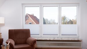 A room with a brown armchair, a wooden table with a metal bucket, and a radiator under large windows.