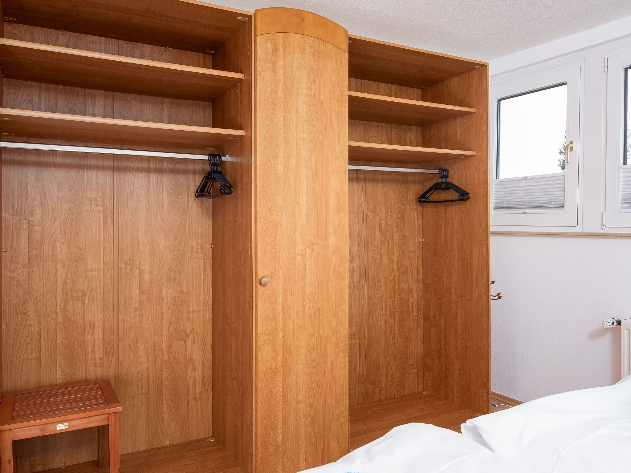 A wooden wardrobe with multiple shelves and hanging space, a small bench, and a window with blinds.