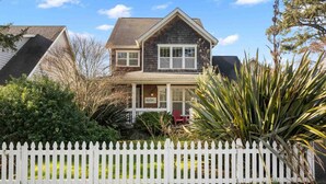 A two-story house with a white picket fence, surrounded by greenery and a clear sky.