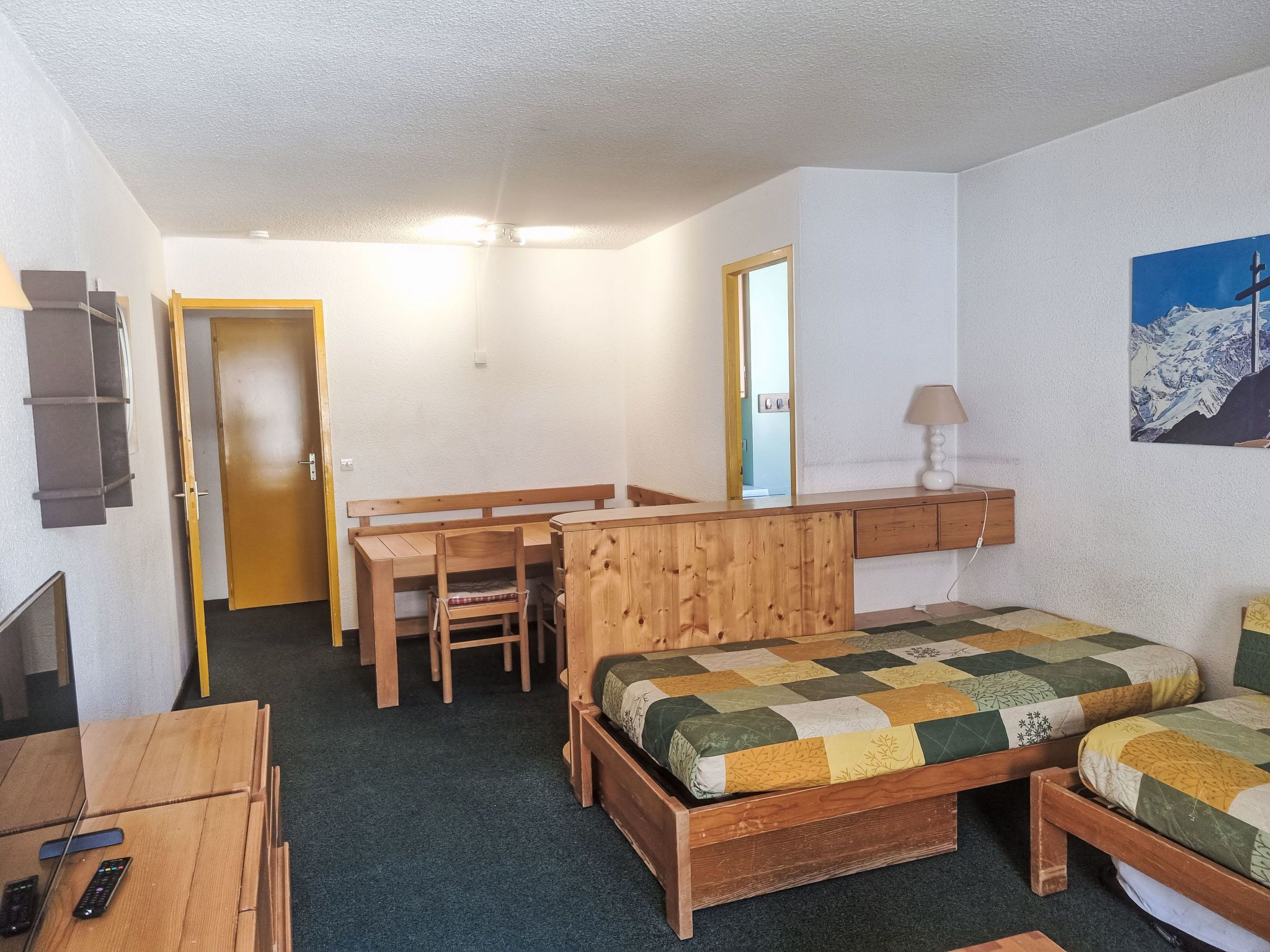 Una habitación pequeña y acogedora con una cama, un escritorio y una silla. Hay una puerta que da a otra habitación, y un cuadro en la pared.