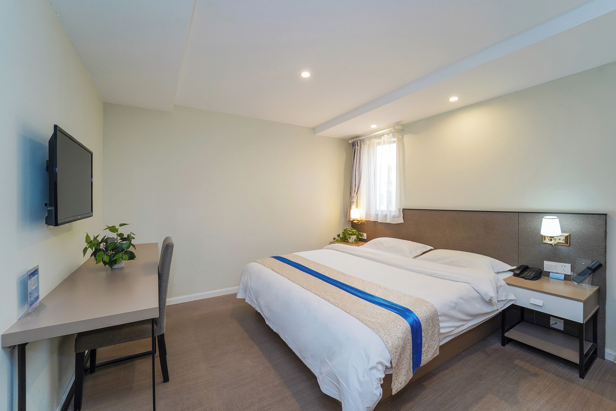 Una habitación de hotel con cama, mesita de noche, escritorio, silla, televisor y ventana con cortinas.