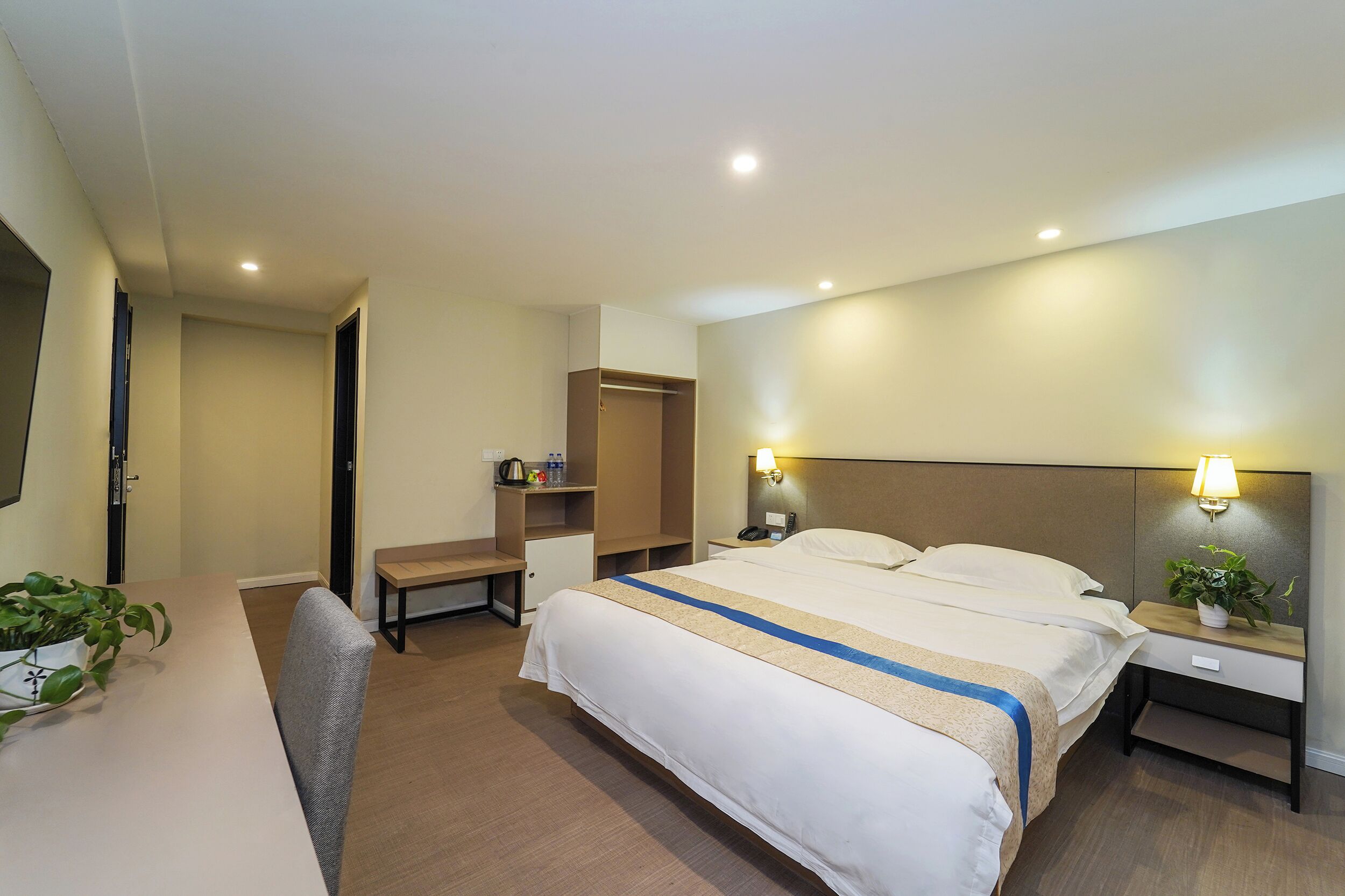 Quarto de hotel com cama, mesas de cabeceira, escrivaninha, cadeira, guarda-roupa e televisão.