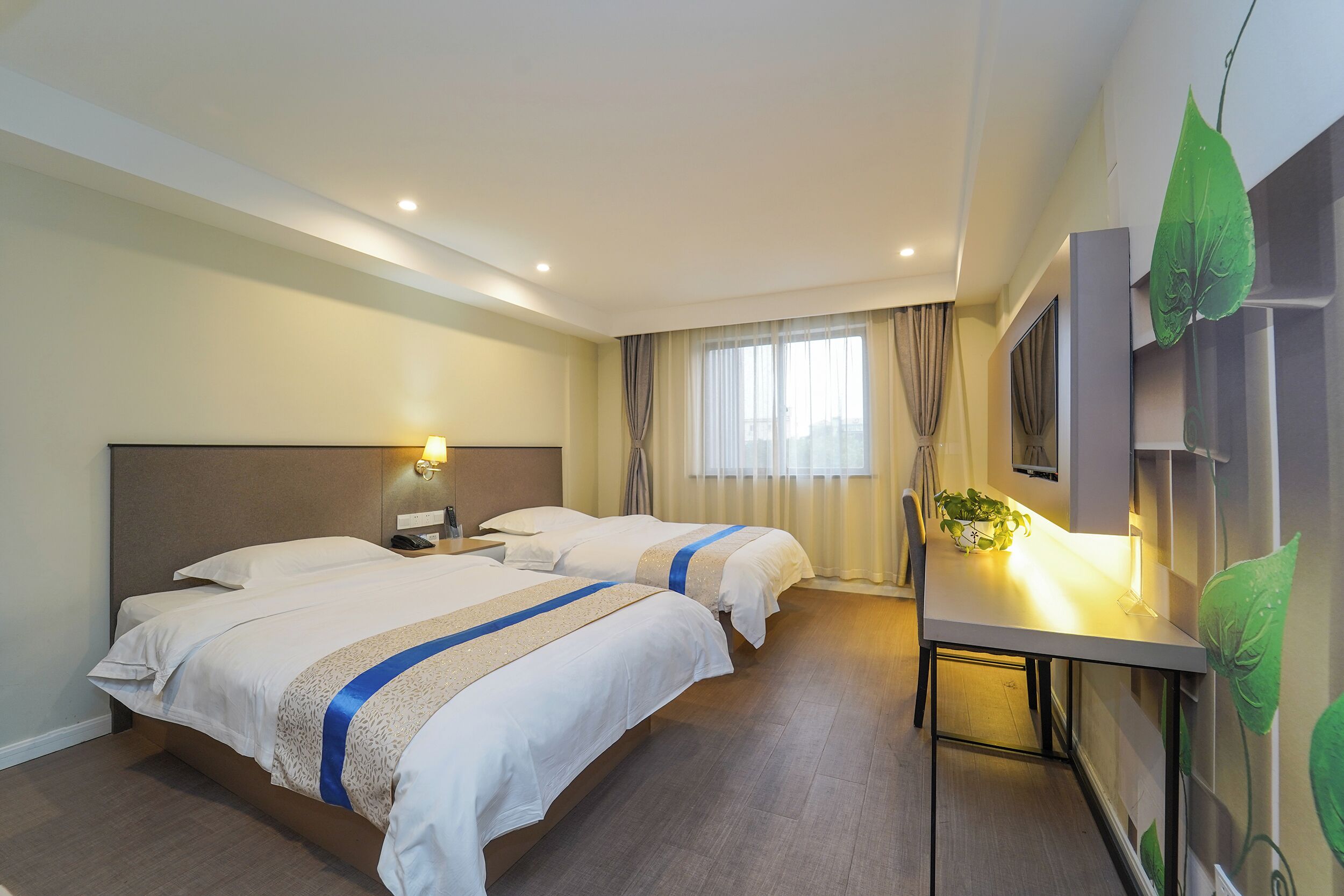 A hotel room with two beds, a desk, a chair, a plant, and a large window with curtains.