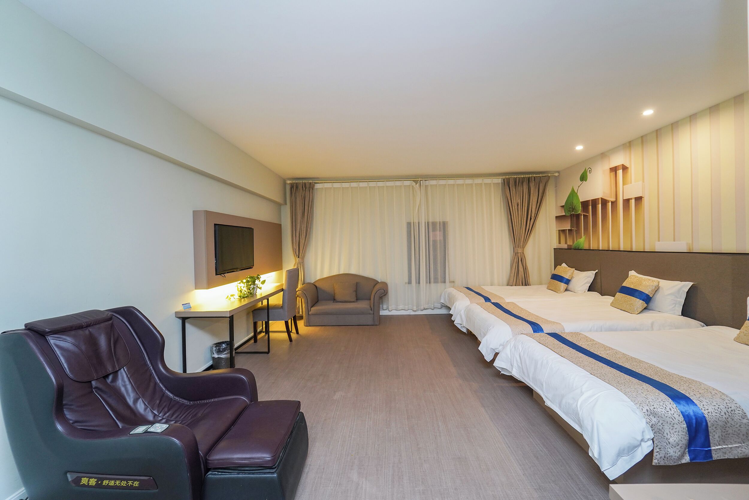 A hotel room with a massage chair, a desk with a lamp, two beds with white bedding and blue accents, a sofa, and a TV on the wall.