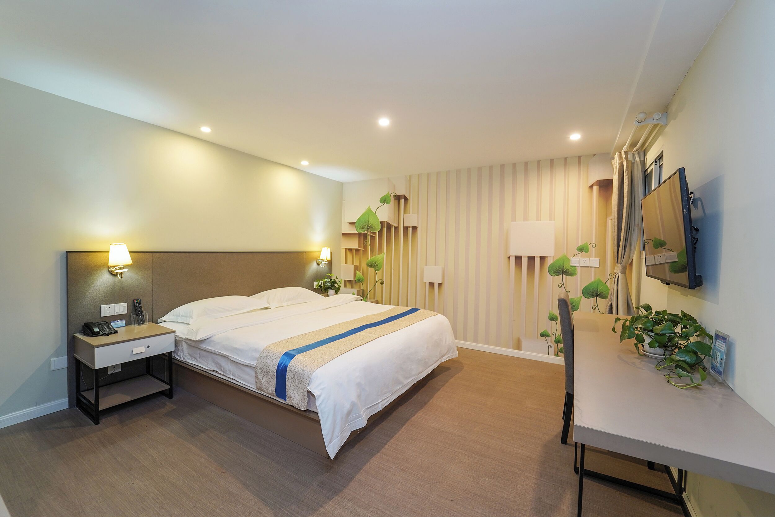 A modern hotel room with a bed, bedside table, lamp, and a desk with a chair and potted plant.
