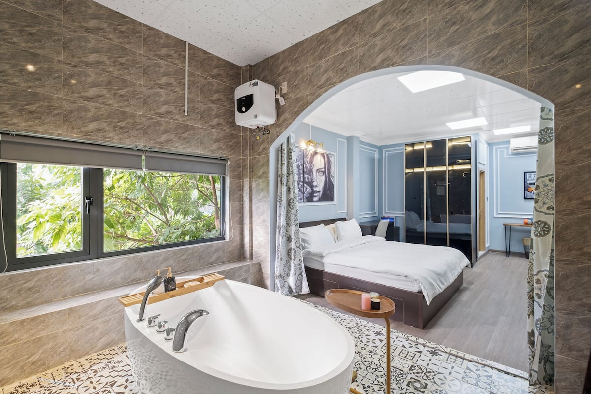 A modern bathroom with a freestanding bathtub, a large window, and a view of a bedroom with a bed and a mirror.