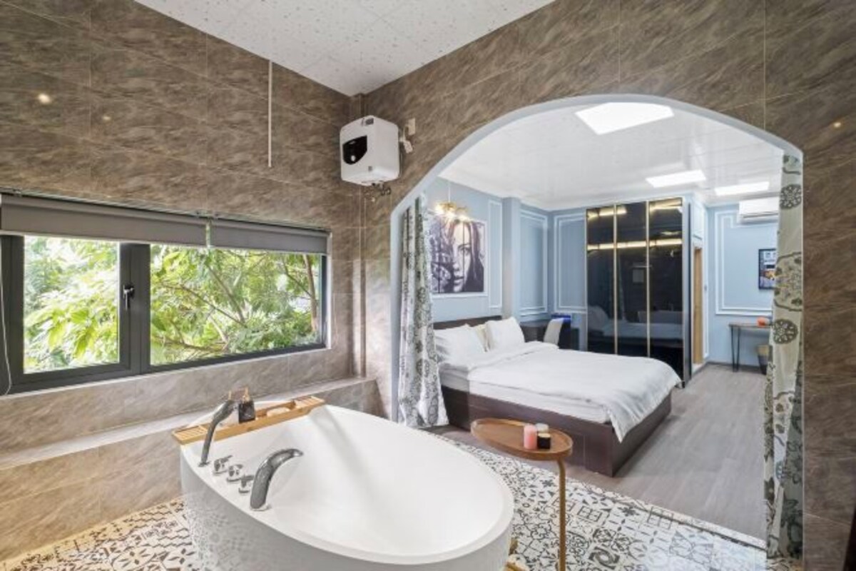 A modern bathroom with a freestanding bathtub, a large window, and a view of a bedroom with a bed and a desk.