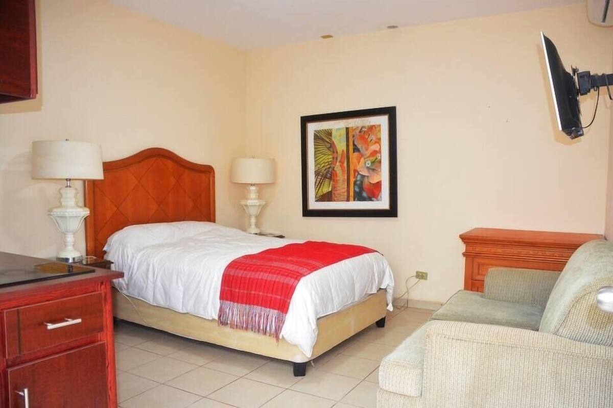 A bedroom with a bed, two bedside lamps, a painting, a television, a desk, and a chair.