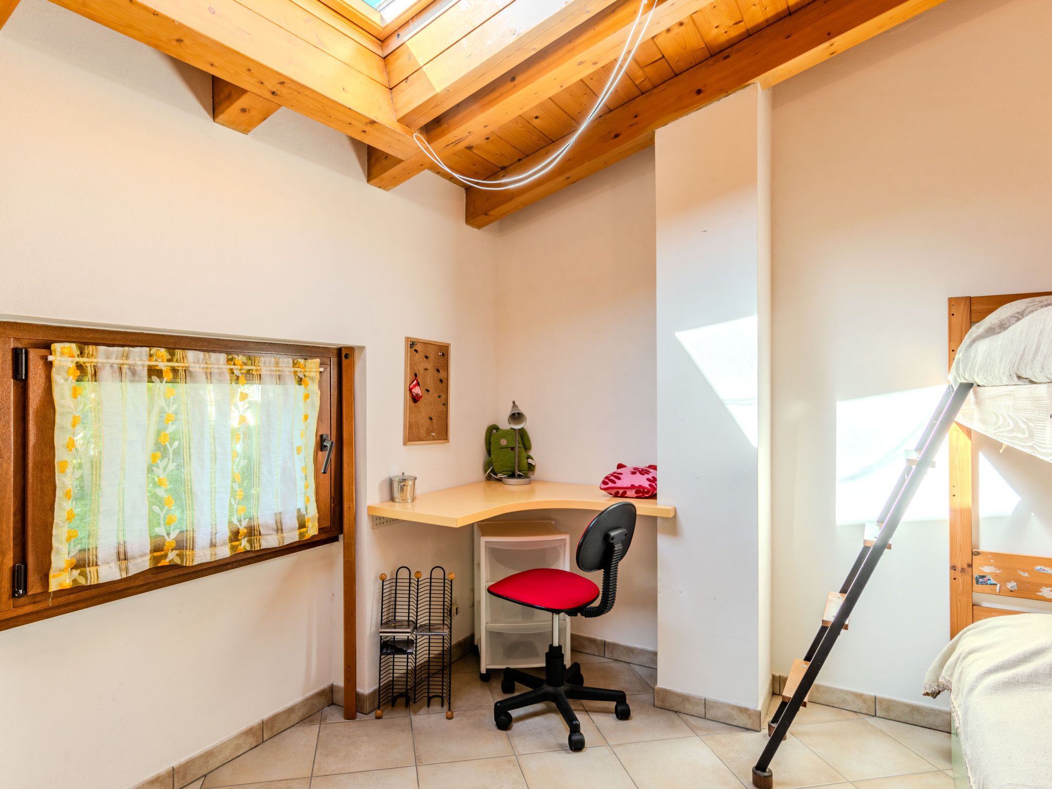 A small, well-lit room with a wooden ceiling, a desk with a chair, a window with curtains, and a bunk bed.