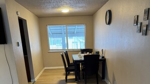 Un comedor con una mesa y sillas, una ventana con persianas, un reloj en la pared y un enchufe en el suelo.