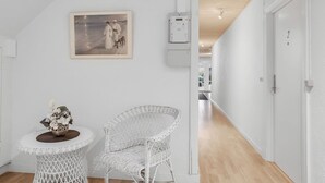 Un pasillo con paredes blancas, techo de madera, un cuadro enmarcado, una silla de mimbre blanca y una pequeña mesa redonda con un florero.